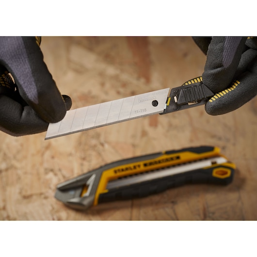 STANLEY 0-11-718 snap-off utility blade held in gloved hands, yellow and black knife in background.