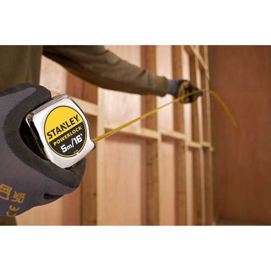 STANLEY 0-33-158 PowerLock tape measure held by gloved hand, measuring wooden wall frame.