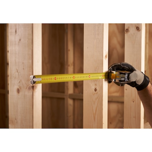 STANLEY 0-33-442 tape measure extended across wooden studs, held by gloved hand in construction setting.