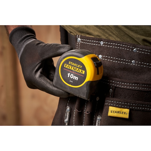 STANLEY 0-33-811 tape measure FATMAX 10m held by gloved hand, clipped to tool belt, close-up view.
