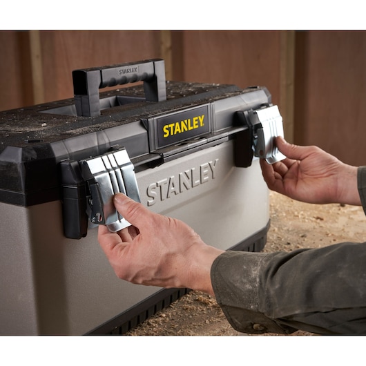 Photo of STANLEY 1-95-616 tool box with metal latches, hands opening the lid in workshop setting.