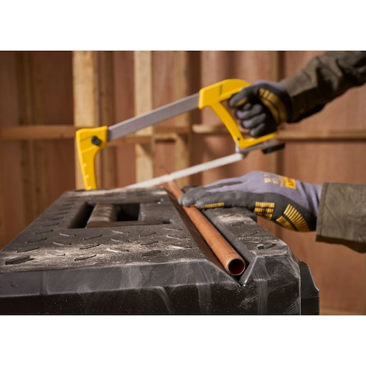 STANLEY 1-95-829 hacksaw held by gloved hands, cutting copper pipe on a black workbench, side angle.