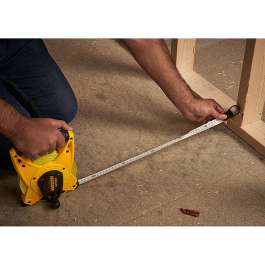 STANLEY 2-34-814 open reel tape measure used on construction floor, yellow case, person measuring wood frame.