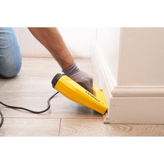 STANLEY 6-TRE550 electric staple gun in use on baseboard, yellow body, black accents, indoor flooring.