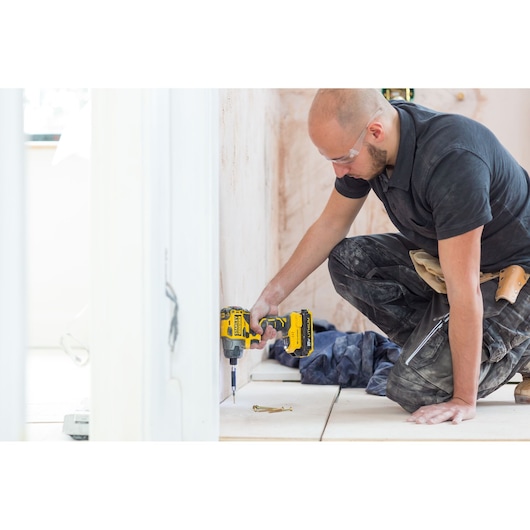STANLEY FMC647D2 cordless drill used by a worker kneeling indoors near a wall, tool bag visible at waist.