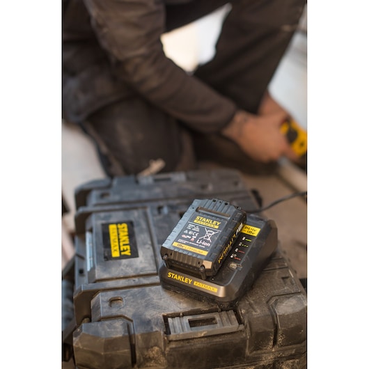 STANLEY FMC692L battery charger on black tool case, close-up with worker using a tool in blurred background.