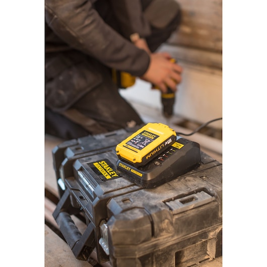 Photo of STANLEY FMC692L battery charger with yellow 18V lithium battery on jobsite toolbox, worker using drill in background.