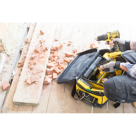 STANLEY FMCB100B cordless drill with yellow tool bag on wooden floor, scattered bricks and dust visible.