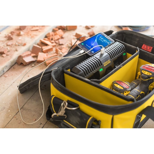 STANLEY FMCB100B battery charger in yellow tool bag charging power tool batteries and a smartphone on a worksite.