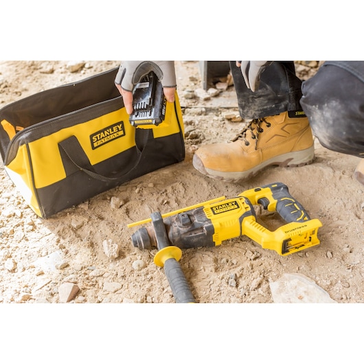 STANLEY FMCD900B cordless rotary hammer drill and yellow-black tool bag on construction site ground.