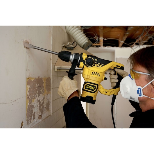 Photo of person using STANLEY FME1250K rotary hammer, drilling into wall, with gloves and safety mask.