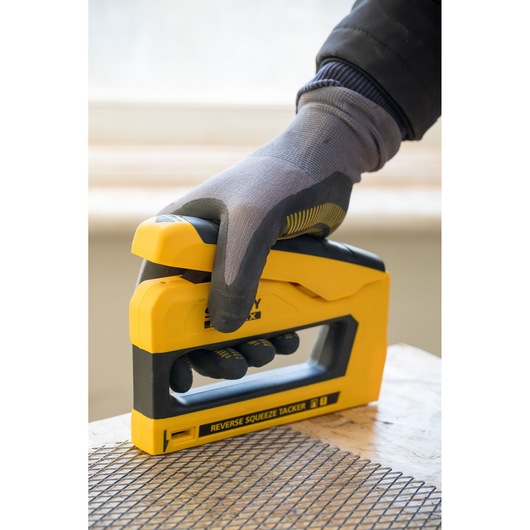 STANLEY FMHT0-80551 tacker in yellow and black, held by a gloved hand, pressing mesh onto wood surface.