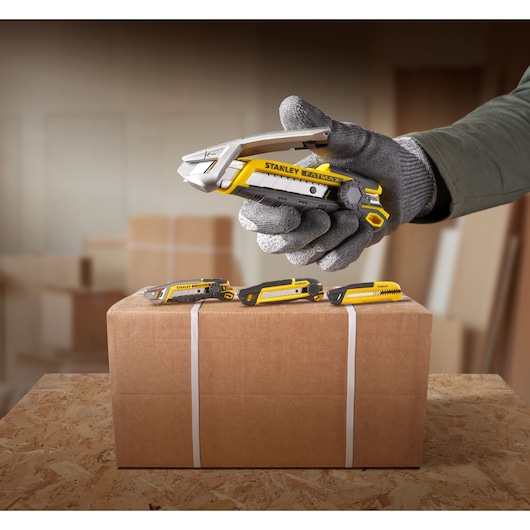 Photo of STANLEY FMHT10592-0 utility knife held in gloved hand above a cardboard box with three knives displayed on top.
