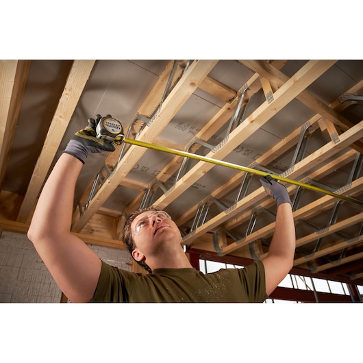 STANLEY FMHT38217-0 tape measure used by person measuring wooden ceiling beams on construction site.