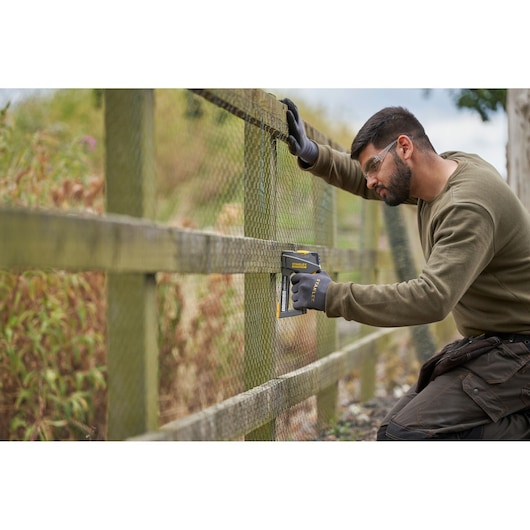 STANLEY FMHT6-70868 stapler used outdoors on mesh fencing, worker wearing gloves, side view.