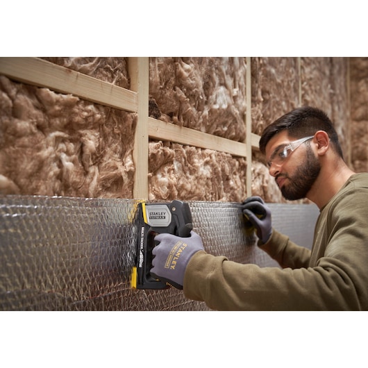 Photo of STANLEY FMHT70401-0 hand stapler being used to secure insulation with gloved hand, construction setting.