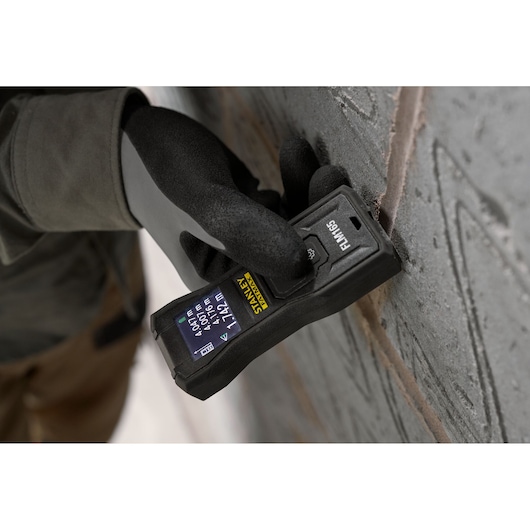 Stanley FMHT77165-0 laser distance measurer held against a wall by a gloved hand, close-up view.