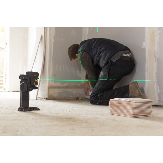 STANLEY FMHT77598-1 laser level projects green lines for wall alignment, worker kneeling nearby, stack of tiles visible.