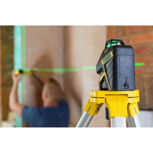 STANLEY FMHT77617-1 green laser level on tripod projecting line across brick wall, worker measuring in background.