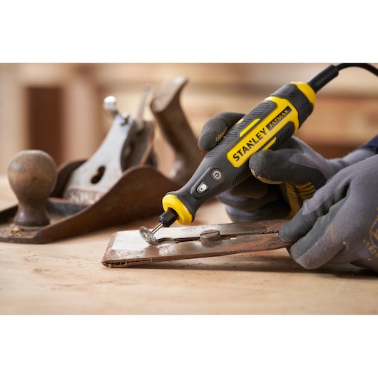 Photo of STANLEY SFMCE030B rotary tool being used to grind metal, handled with gloves, woodworking tools nearby.