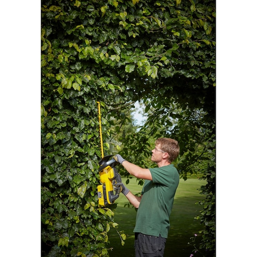 STANLEY SFMCHTB866B hedge trimmer in use outdoors, yellow and black tool cutting thick green hedge.