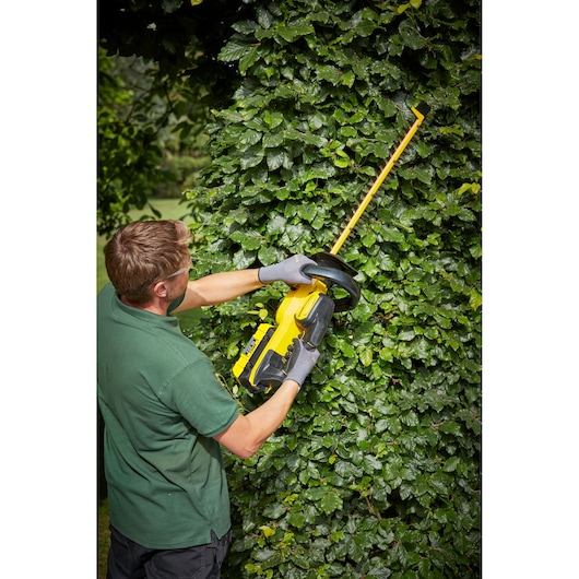 Photo of a person using a STANLEY SFMCHTB866B hedge trimmer outdoors, trimming dense green bushes.