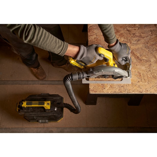 Top view of a person using a STANLEY SFMCS550B cordless circular saw to cut wood, attached to a dust extractor.