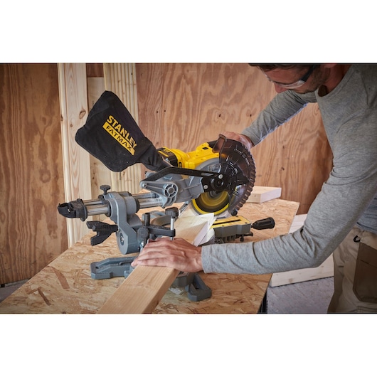 STANLEY SFMCS701M1 miter saw in use cutting wood on a workbench, dust bag attached, indoor construction setting.