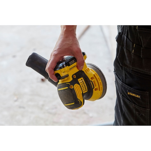 Photo of STANLEY SFMCW220B cordless orbital sander in hand, yellow and black body, work pants visible.