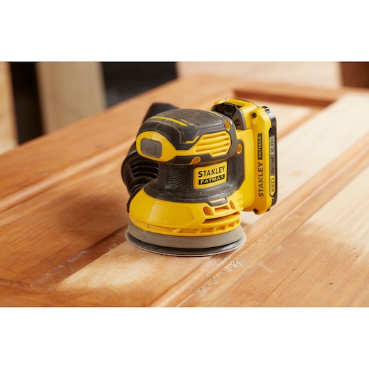 STANLEY SFMCW220D1S cordless orbital sander in yellow and black, sanding a wooden surface.