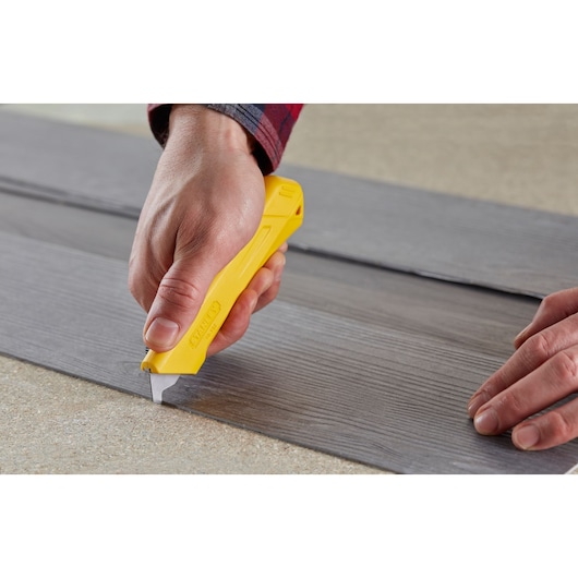 STANLEY STHT0-10192 yellow utility knife cutting grey vinyl plank flooring, close-up side view.