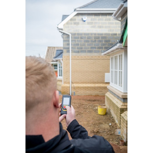 Photo of STANLEY STHT1-77140 laser distance measurer held by a person outdoors near a construction site.