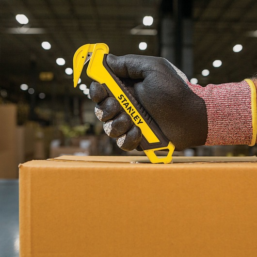 Stanley STHT10356 box cutter held with glove, cutting cardboard box in warehouse setting.