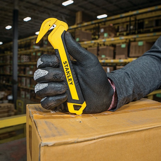 Photo of STANLEY STHT10356 yellow safety box cutter in gloved hand opening cardboard box in warehouse.