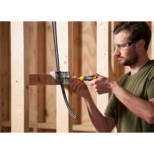 Photo of person using STANLEY STHT16163-0 screwdriver on an electrical box, wooden frame background.