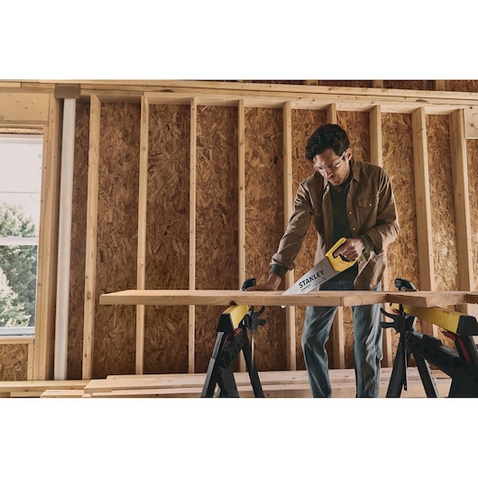 STANLEY STHT20348 hand saw used to cut wood plank on sawhorses inside unfinished building.