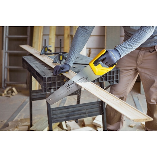 Stanley STHT20349-1 hand saw with yellow handle cutting wood plank on workbench, close-up angle.
