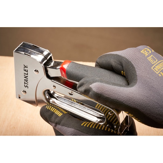 STANLEY STHT6-70410 chrome staple gun held in gloved hands, side angle on wooden surface.