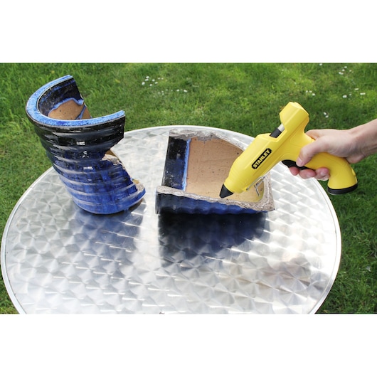 Photo of a STANLEY STHT6-70416 glue gun being used to repair a broken blue ceramic pot on a metal table outdoors.