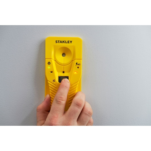 STANLEY STHT77587-0 yellow stud finder held against wall, person pressing button, front view.