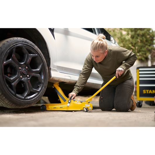 STANLEY STMT81251-1 yellow car jack in use, side angle, with person and tool chest nearby outdoors.