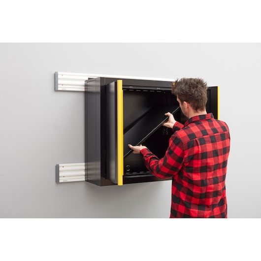 STANLEY STST97598-1 wall-mounted black storage cabinet with yellow trim, person inserting shelf, side view.